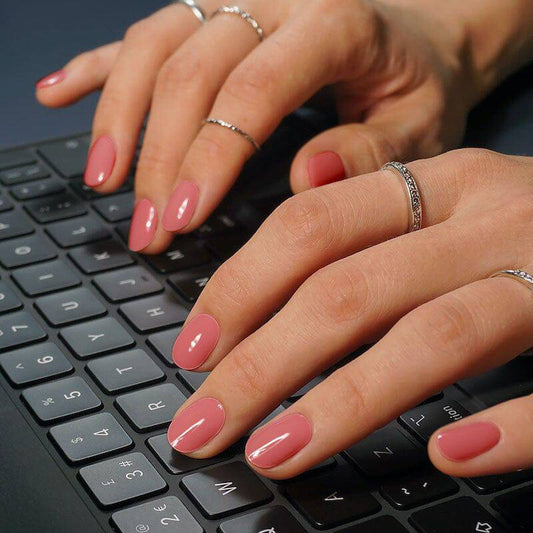 Manicured nails painted in a warm, rosy pink polish with a glossy finish. The hands are positioned over a keyboard, suggesting a work or tech setting.
