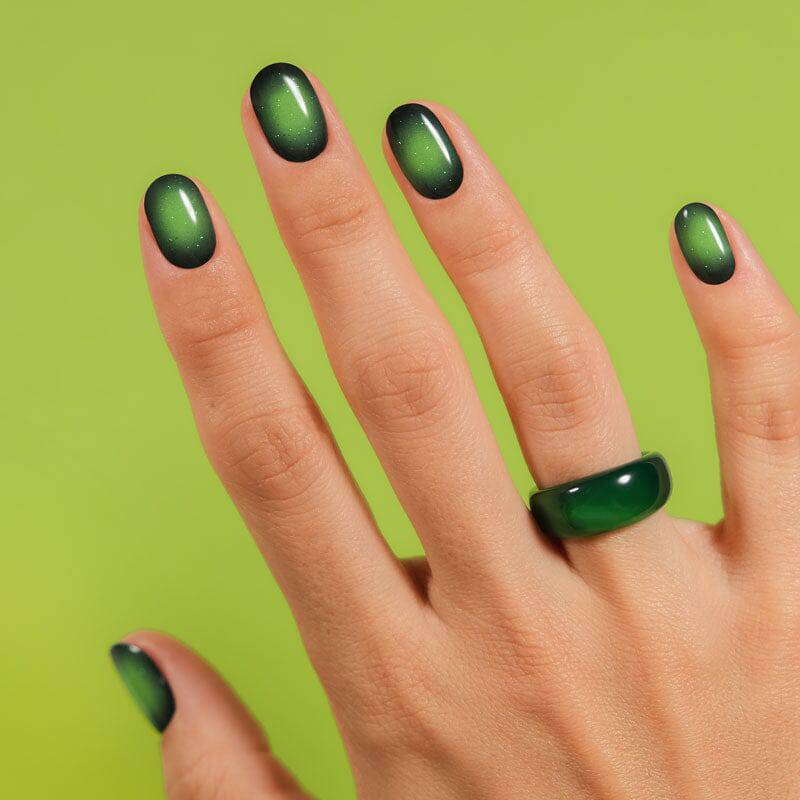 A hand with nails painted in a shimmering green, creating a gradient effect. The background features a bright green surface, enhancing the vibrant design.
