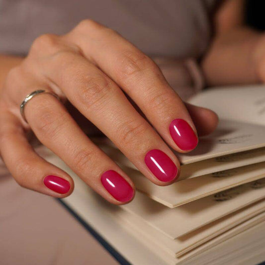 A close-up of a hand with nails painted in a deep fuchsia polish, with a glossy finish. The hand is resting on a stack of cream-colored papers.