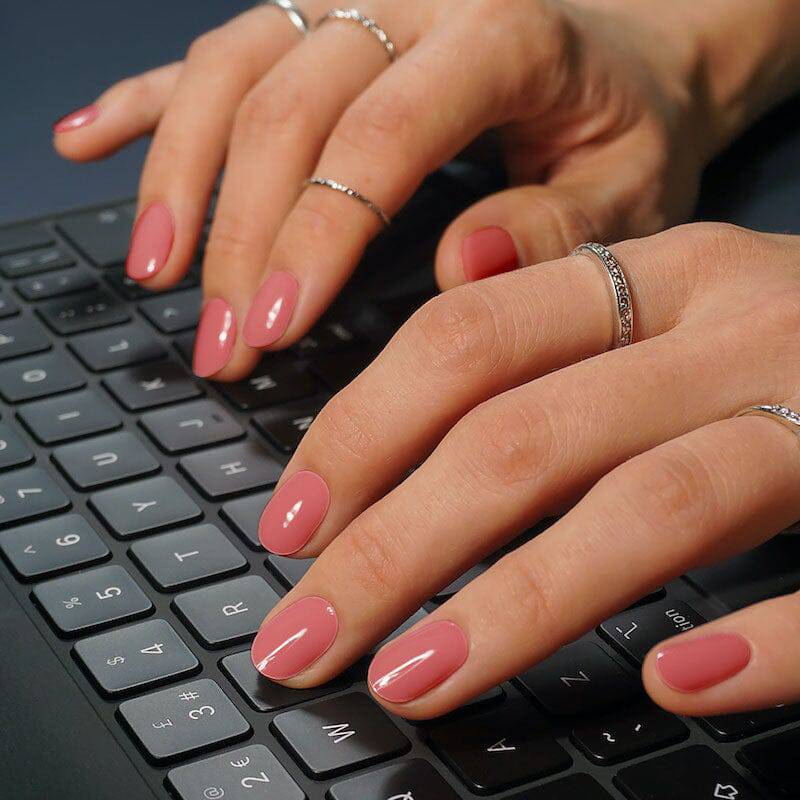 Manicured nails painted in a warm, rosy pink polish with a glossy finish. The hands are positioned over a keyboard, suggesting a work or tech setting.