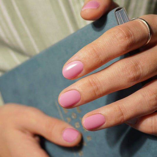 Nails painted in a soft, glossy pink polish. The hand holds a blue book, creating a casual and artistic composition.