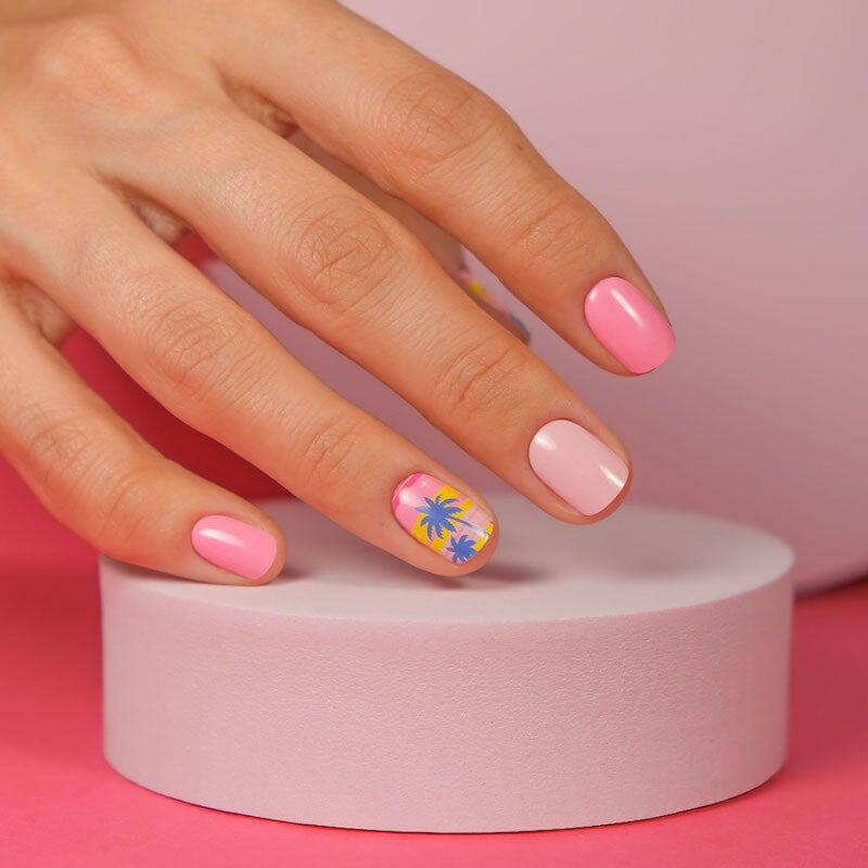 Nails painted in shades of pink with one accent nail featuring tropical palm tree art. The background includes soft pink and neutral tones.