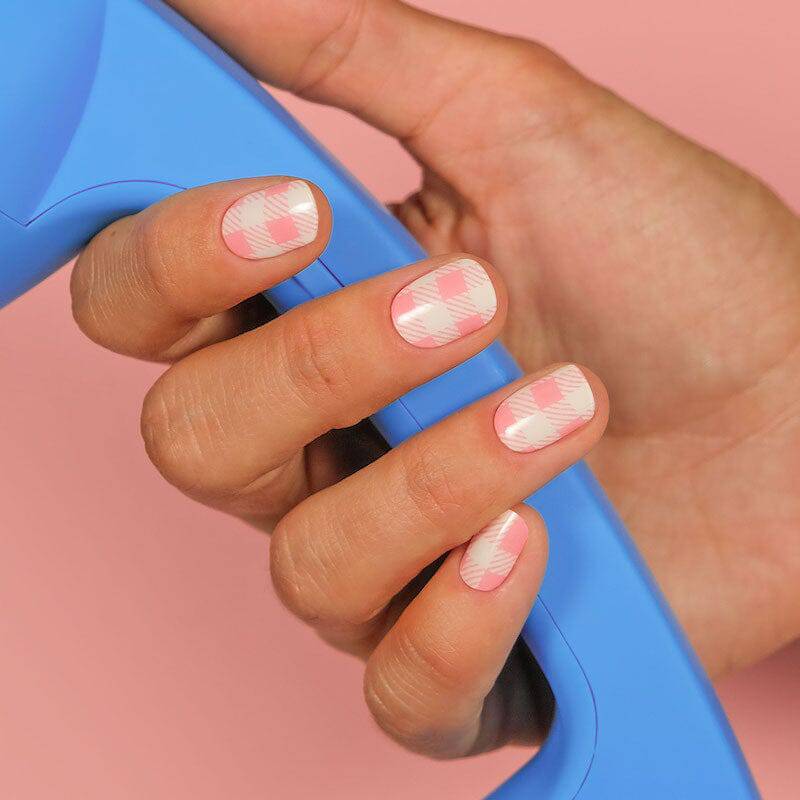  Close-up of nails featuring pink and white checkered patterns. The hand is holding a blue item against a pastel pink background.
